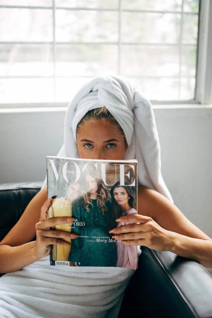 Woman wrapped in towel reading Vogue magazine in a relaxed indoor setting.